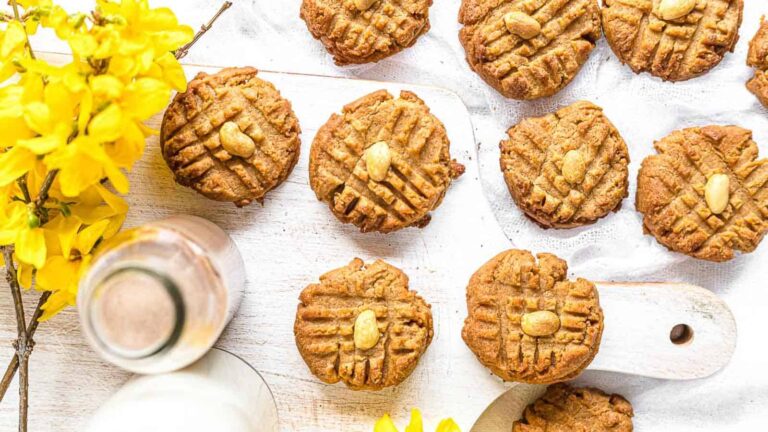 Peanut butter cookies with peanut toppings arranged on a white wooden board, next to a glass bottle of milk and yellow flowers.