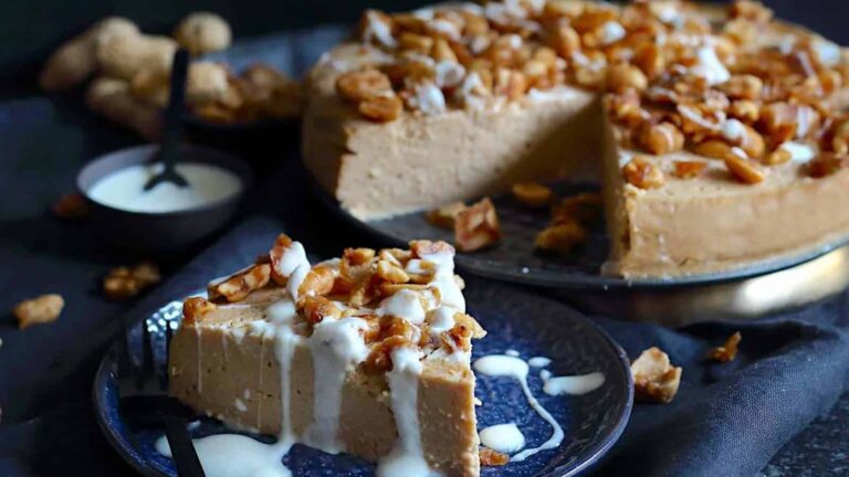 A slice of creamy nut-topped cheesecake is served on a plate with a drizzle of white sauce, next to a larger cheesecake with a cut section.