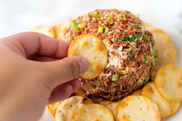 A hand holding a round cracker, reaching to dip into a pecan bacon cheeseball.