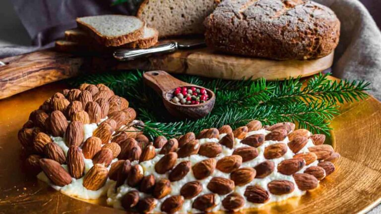 A cheese ball shaped like a hedgehog, decorated with almonds, is displayed on a wooden board next to sliced brown bread and a small spoon of mixed peppercorns.