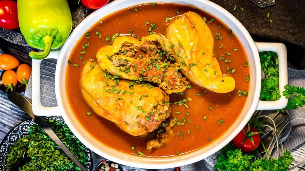 Stuffed yellow bell peppers filled with ground meat and rice sit in a tomato-based sauce, garnished with chopped herbs in a white casserole dish.