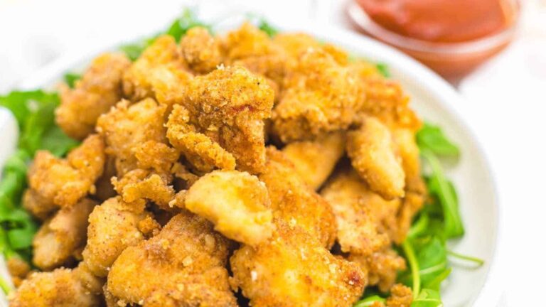 A bowl of crispy, golden-brown breaded chicken pieces served on a bed of greens, with a small dish of red dipping sauce in the background.
