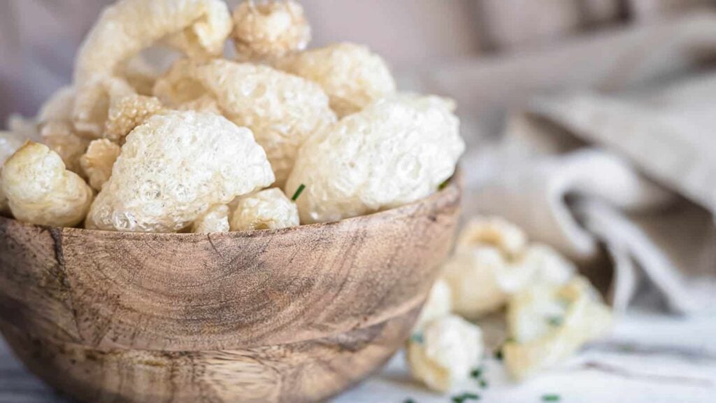 A wooden bowl filled with light, crispy pork rinds sits on a marble surface, with a few pieces scattered nearby.