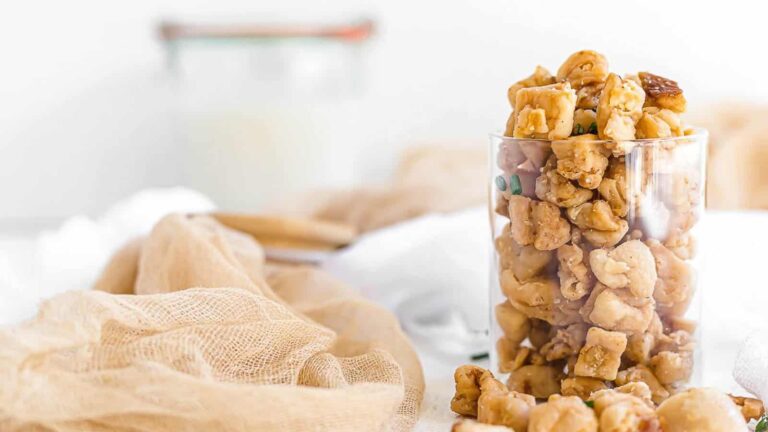 A clear glass filled with caramel-coated snack clusters sits on a light-colored surface with a jar of milk and gauzy fabric in the background.