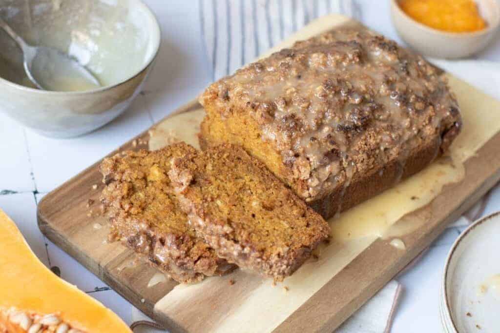 A loaf of glazed pumpkin bread with two slices cut sits on a wooden cutting board, surrounded by bowls containing glaze and orange puree.