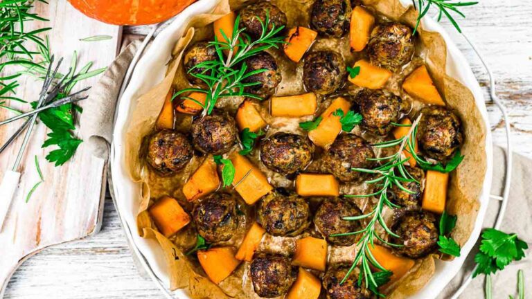 A round baking dish lined with parchment paper contains cooked meatballs, cubed squash, sprigs of rosemary, and fresh parsley.