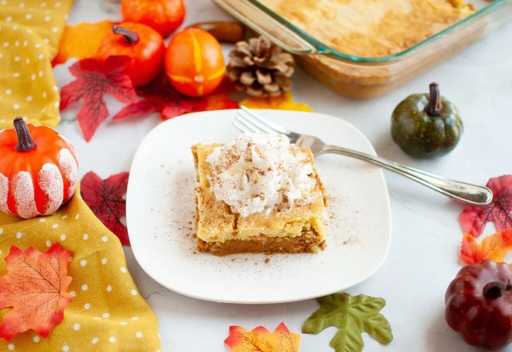 A slice of pumpkin dessert with whipped cream on a white plate, surrounded by autumn decorations like leaves and small pumpkins.