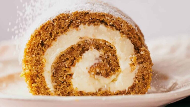 A close-up of a pumpkin roll cake with cream cheese filling, dusted with powdered sugar, on a white plate.