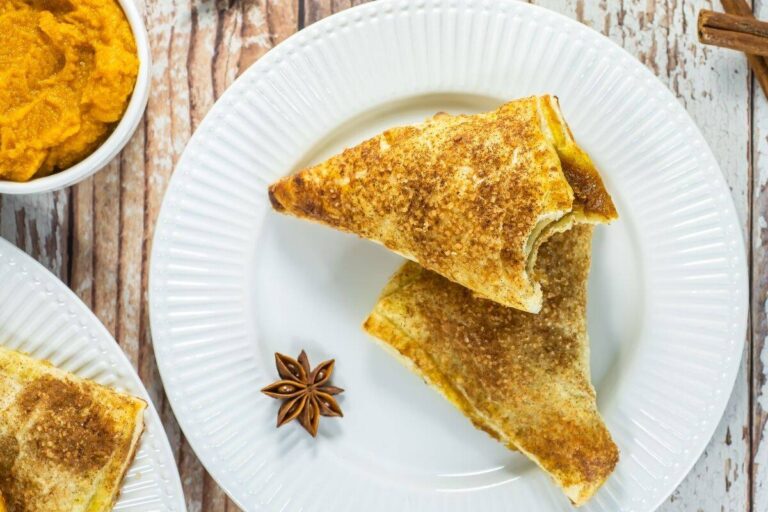 Two triangular pastries with a golden-brown crust are placed on a white plate, next to a star anise, on a wooden table. A bowl of orange filling is partially visible.