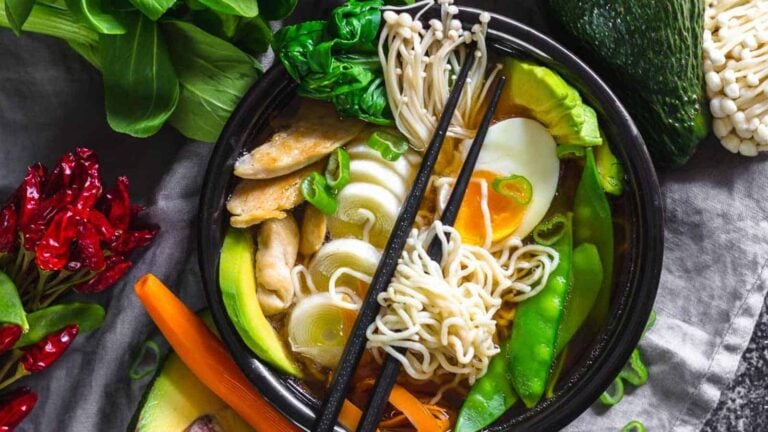 A bowl of ramen with noodles, sliced boiled egg, vegetables, mushrooms, and chicken, garnished with green onions, surrounded by fresh produce.