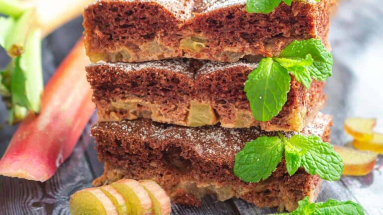 Three slices of rhubarb cake stacked on a wooden surface, topped with powdered sugar and garnished with fresh mint leaves and rhubarb pieces.
