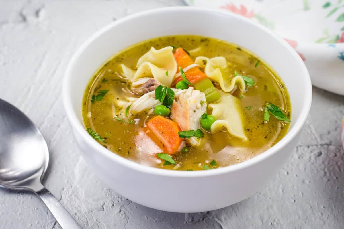 A bowl of Rotisserie Chicken Noodle Soup topped with parsley on a white surface, next to a spoon.