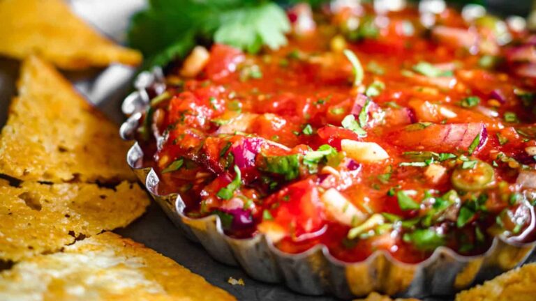 A close-up of a metal dish filled with chunky salsa, garnished with herbs, next to several triangular tortilla chips.