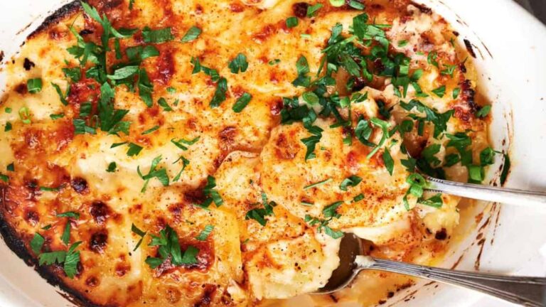 A baked dish of cheesy scalloped potatoes topped with chopped parsley, with a serving spoon partially lifting a portion.