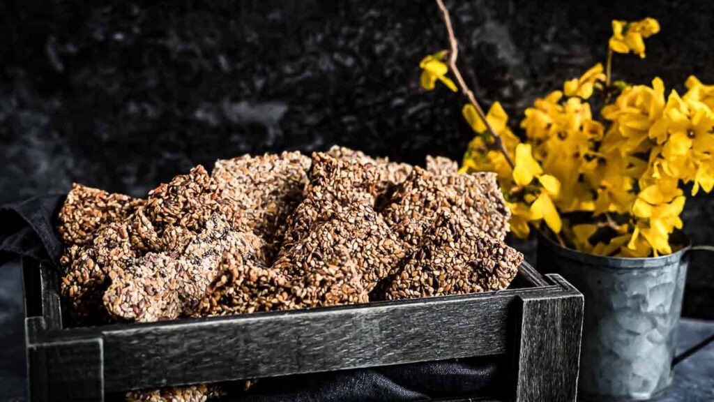 A black wooden tray filled with seed crackers sits next to a small metal bucket with yellow flowers against a dark background.