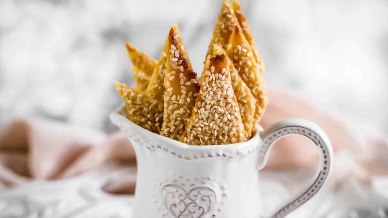 Sesame-covered pastry triangles arranged upright in a decorative white ceramic pitcher with a heart design.