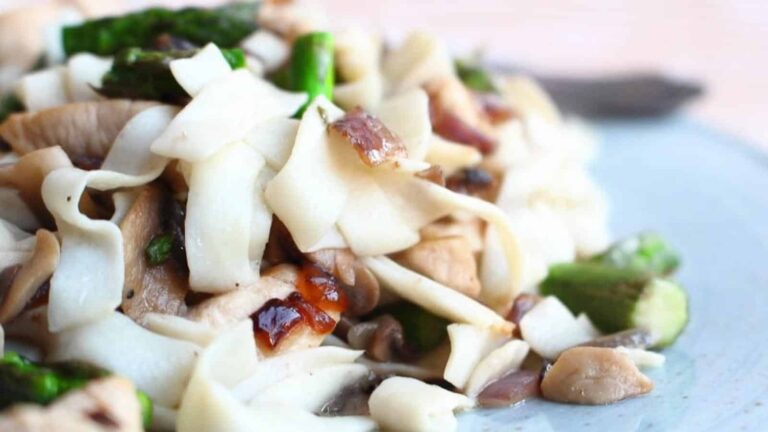 A plate of wide rice noodles mixed with pieces of chicken, asparagus, mushrooms, and onions.