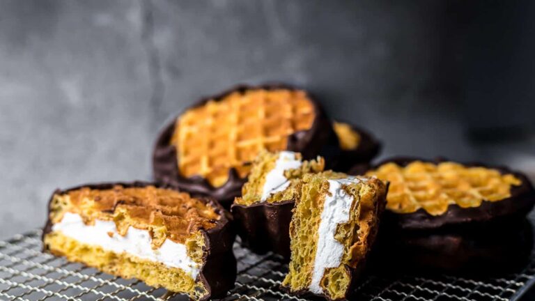 Chocolate-covered waffle sandwiches with a marshmallow filling are stacked on a cooling rack, with some cut in half to show the filling.