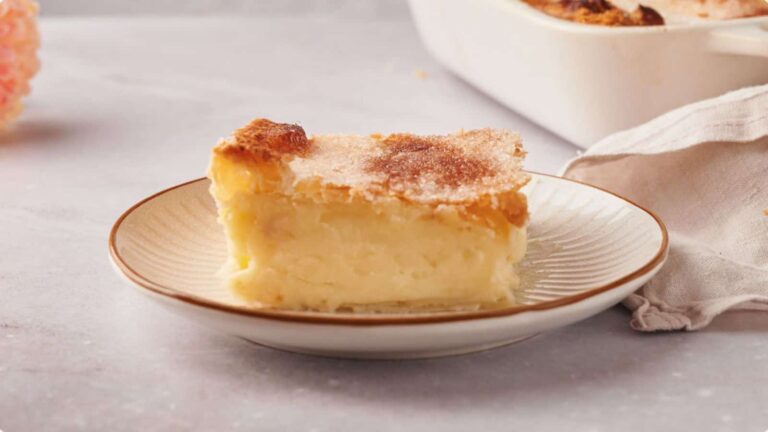 A slice of custard pie with a golden-brown, sugared crust sits on a white plate; a baking dish and napkin are in the background.