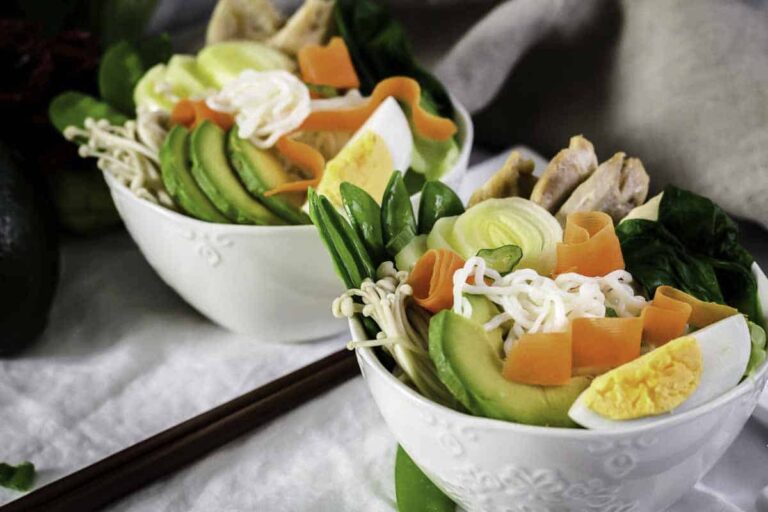 Two white bowls filled with ramen noodles, sliced avocado, boiled egg wedges, carrots, mushrooms, leafy greens, and chicken, with a pair of chopsticks beside them.