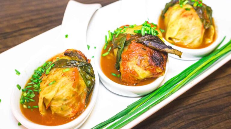 Three stuffed cabbage rolls in sauce are served in individual white dishes, garnished with chopped chives, on a rectangular plate with whole chives on the side.