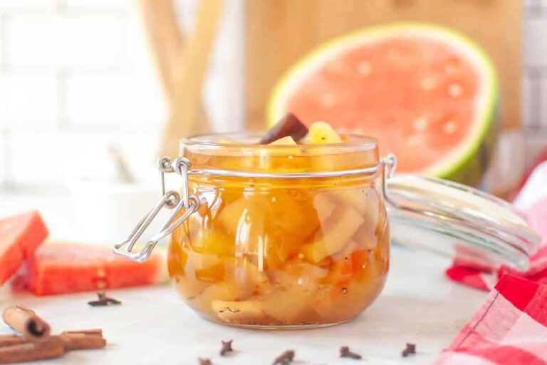A glass jar filled with watermelon rind preserves sits on a table, surrounded by cloves, cinnamon sticks, and sliced watermelon in the background.