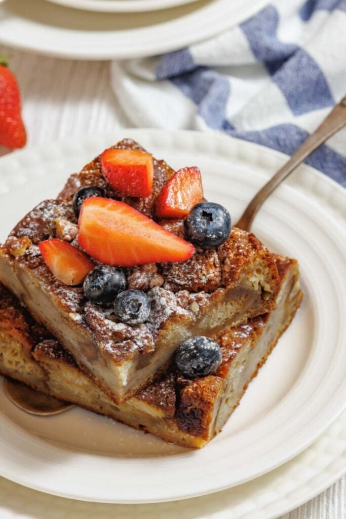 Two slices of bread pudding topped with powdered sugar, fresh strawberries, and blueberries on a white plate with a spoon.