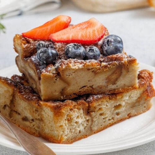 Two slices of bread pudding stacked on a white plate, topped with blueberries and sliced strawberries, with a fork beside them.