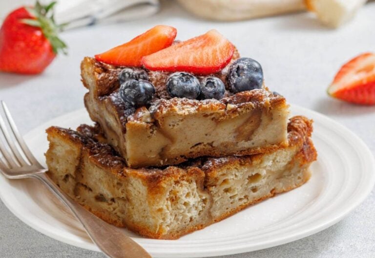 Two slices of bread pudding stacked on a white plate, topped with blueberries and sliced strawberries, with a fork beside them.