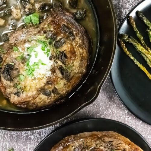 A cooked Broiled Prime Rib Steak topped with herbs and a dollop of butter sits in a cast iron skillet next to a plate of roasted asparagus spears.