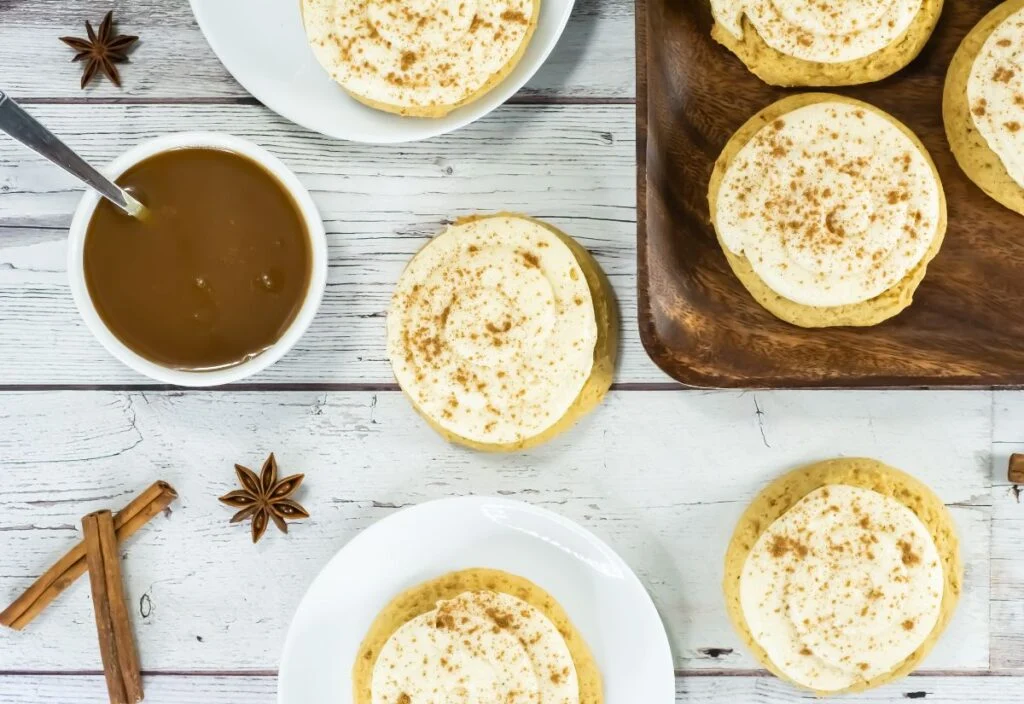 Frosted cookies sprinkled with cinnamon are arranged on white plates and a wooden tray, with a bowl of caramel sauce, cinnamon sticks, and star anise on a white wooden surface.