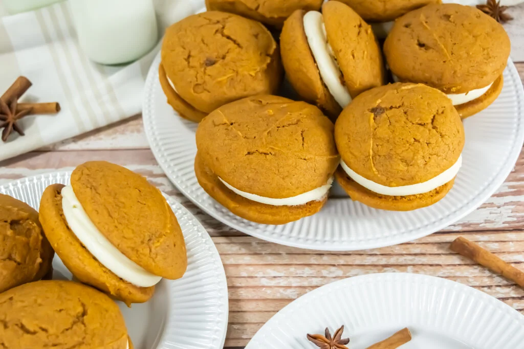 Several pumpkin whoopie pies with cream filling are arranged on white plates, with cinnamon sticks and star anise scattered on a wooden surface.