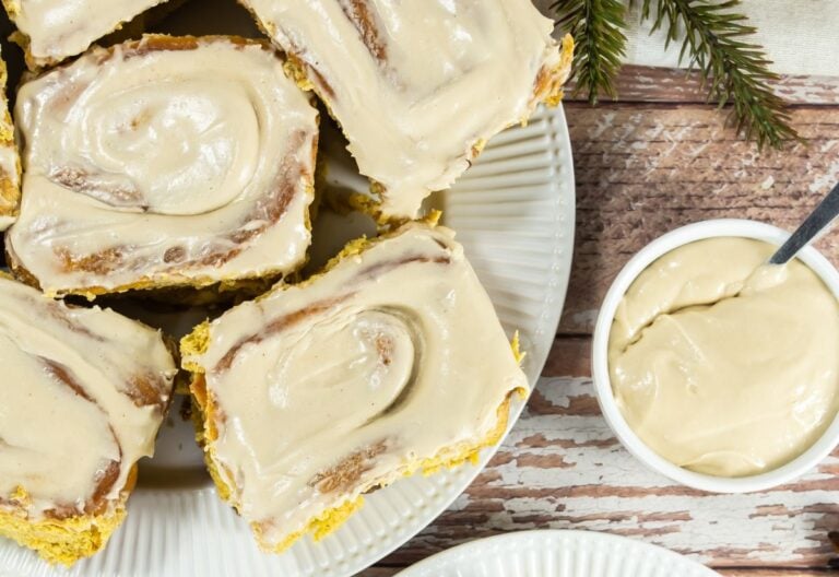 Several frosted cinnamon rolls on a white plate with a small bowl of icing and a spoon beside them, set on a rustic wooden table.