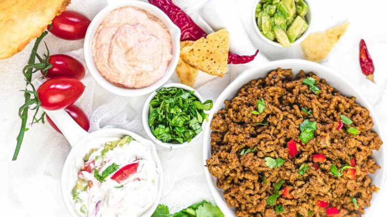 A bowl of seasoned ground meat garnished with cilantro, surrounded by chopped herbs, dips, sliced tomatoes, guacamole, tortilla chips, and red chili peppers.