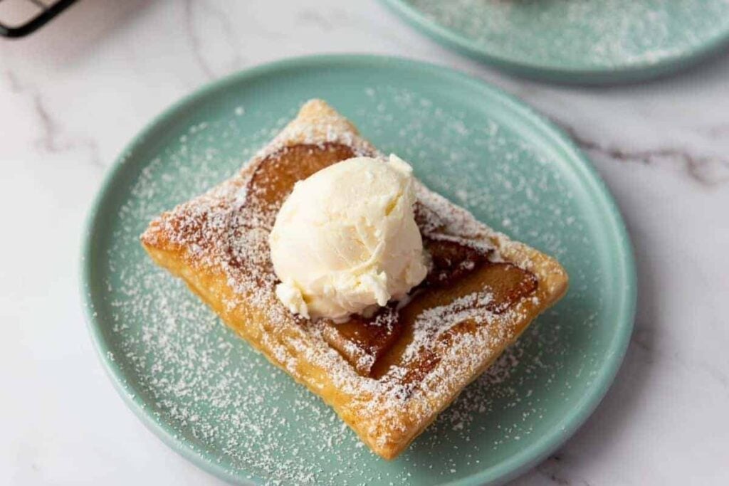 A square pastry dusted with powdered sugar, topped with a scoop of vanilla ice cream, served on a light blue plate.