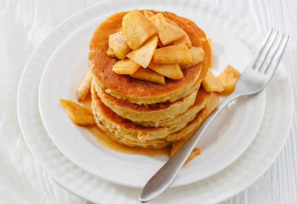 A stack of pancakes topped with cooked apple slices and syrup, served on a white plate with a fork.