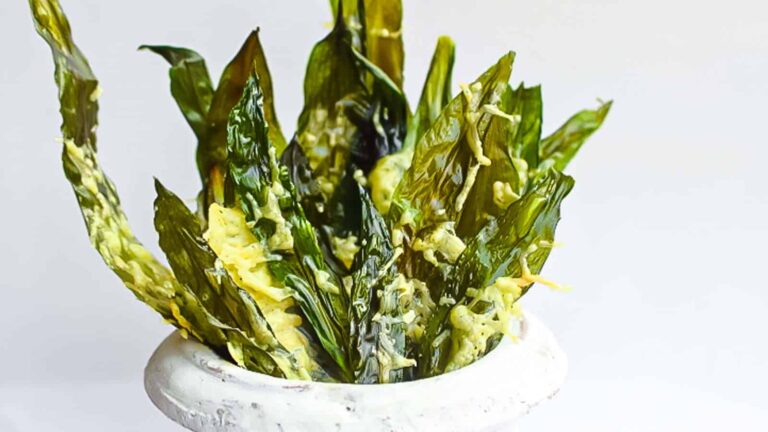 Green wild garlic chips coated with a yellow seasoning are arranged upright in a white bowl against a plain white background.