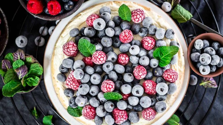 A cheesecake topped with fresh blueberries, raspberries, and mint leaves, surrounded by small bowls of berries and mint on a dark surface.