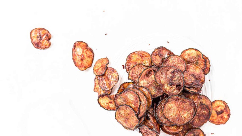 A pile of crispy, round zucchini chips on a white surface, with a few chips scattered to the side.