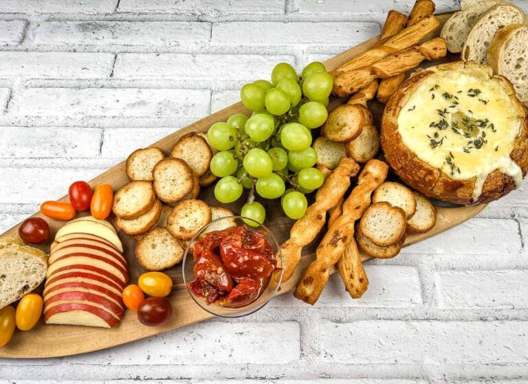 A wooden platter with grapes, sliced apples, cherry tomatoes, toasted bread slices, breadsticks, roasted tomatoes, and a round baked cheese topped with herbs.
