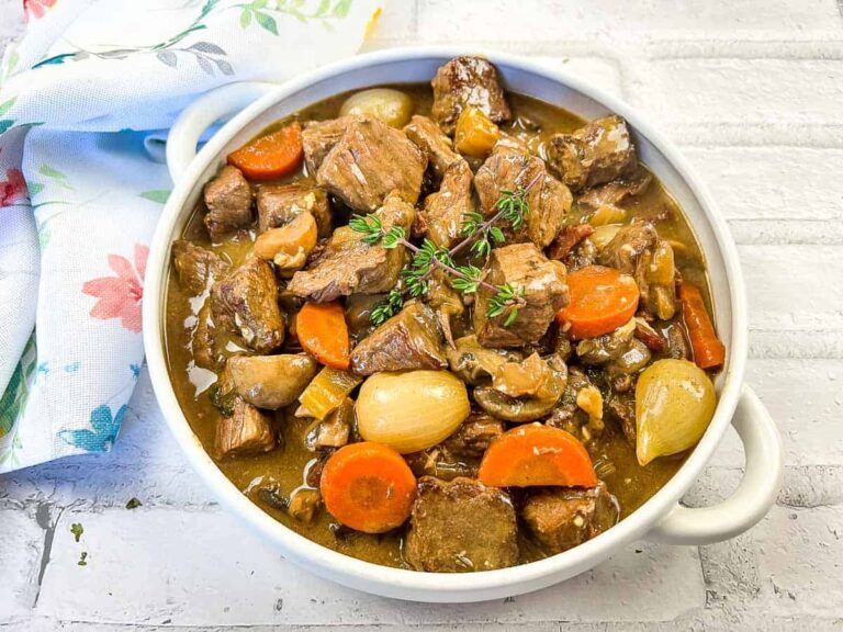 A bowl of beef stew with chunks of beef, carrots, pearl onions, mushrooms, and a sprig of thyme on top, placed on a white surface next to a floral napkin.