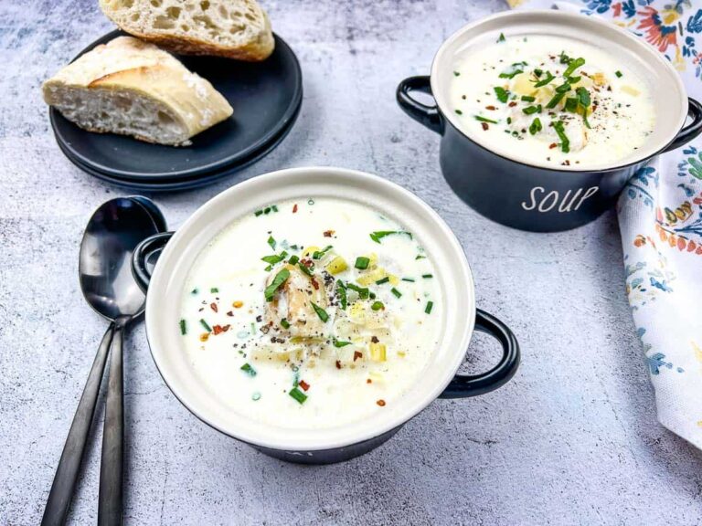 Two bowls of creamy soup topped with herbs and pepper, served with sliced bread on a plate and two spoons on the side.