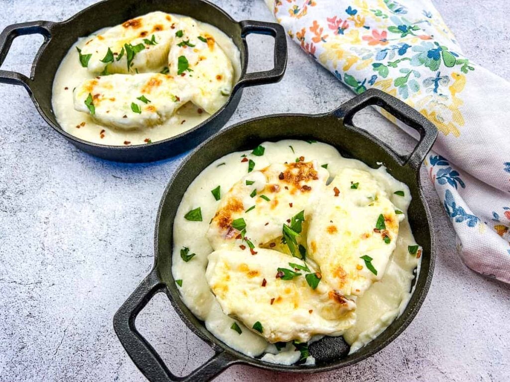 Two small cast iron skillets filled with baked cheese and white sauce, garnished with chopped herbs, on a light textured surface next to a floral-patterned cloth napkin.
