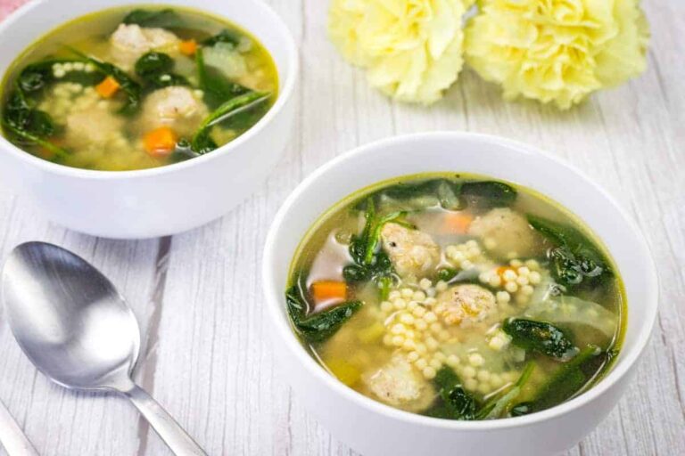 Two white bowls of Italian wedding soup with meatballs, spinach, pasta, and carrots on a light wooden table, next to a spoon and two yellow flowers.