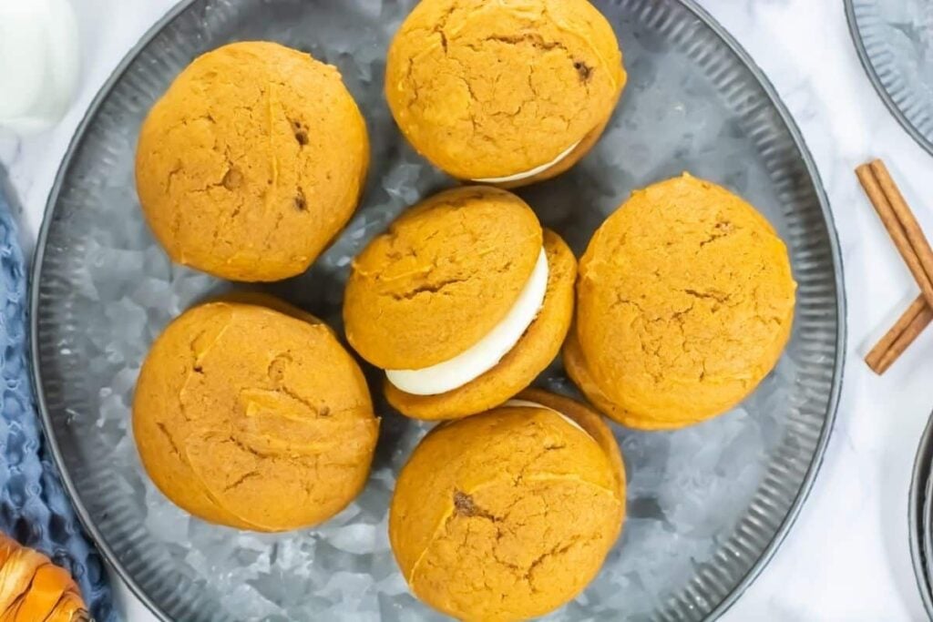 Six pumpkin whoopie pies with cream filling are arranged on a gray plate, viewed from above. Two cinnamon sticks are visible at the edge of the plate.
