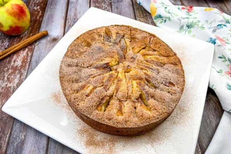 A round apple cake topped with cinnamon sugar is displayed on a white square plate, with an apple, a cinnamon stick, and a floral cloth in the background.