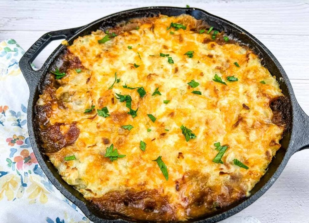 A baked casserole with a golden-brown, melted cheese topping is served in a black cast iron skillet and garnished with chopped parsley.