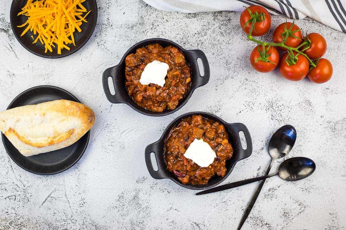 Smoked brisket chili served in two black bowls topped with sour cream, surrounded by fresh tomatoes, shredded cheese, crusty bread, and two spoons on a gray surface.