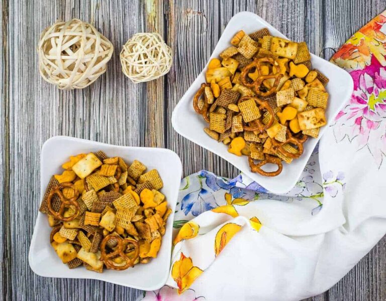 Two bowls of snack mix with pretzels, crackers, and cereal on a wooden surface next to woven decorative balls and a floral-patterned cloth.