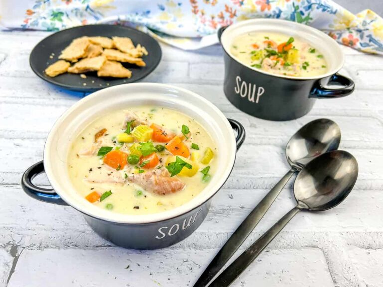 Two bowls of creamy soup with vegetables and meat, garnished with green onions, served with crackers on a black plate and two spoons on a white surface.